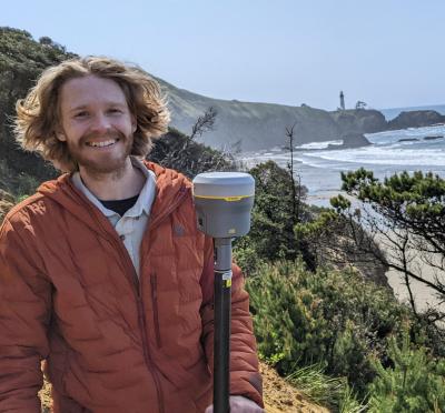 A young man poses along a coastal shoreline holding a piece of surveying equipment.