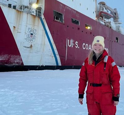 A woman in cold weather gear poses in a snowy landscape in front of a research vessel.