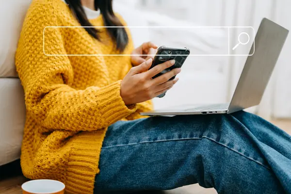 A person sits on the floor with a laptop on their lap and a smartphone in their hand, using both simultaneously. A search bar is superimposed over the image.