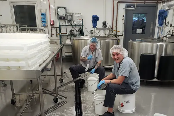 Two students in hairnets and gloves smile at the camera while working in a commercial cheese-making facility.