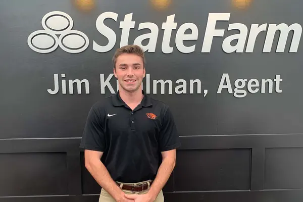 Adam Henderson, wearing an OSU Beaver polo shirt and khakis, poses in front of a State Farm sign at his internship.