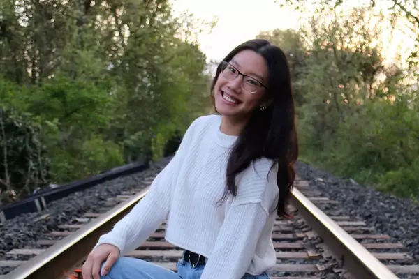 Anna Lane, wearing a white sweater and blue jeans, smiles at the camera, posing in an outdoor setting