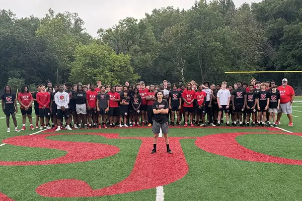 Brenn Hardaway, wearing athletic shorts and a T-shirt, poses on a sports field in front of a crowd of youth athletes