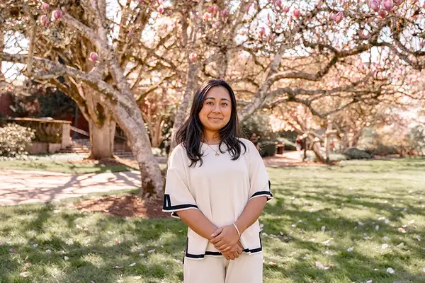 Hana Nakamura-Daya, wearing a white blouse and pants, poses in front of a green lawn and flowering tree on the OSU campus.