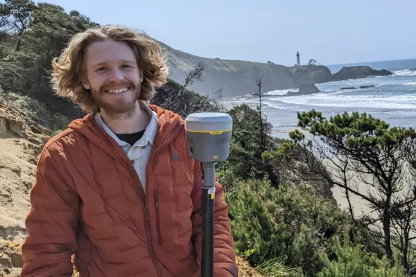 A young man poses along a coastal shoreline holding a piece of surveying equipment.