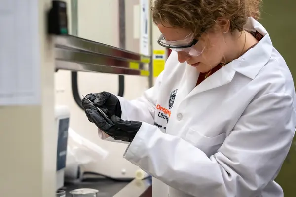 Madalyn Gragg, wearing a white lab coat, safety goggles and gloves, works in a science lab