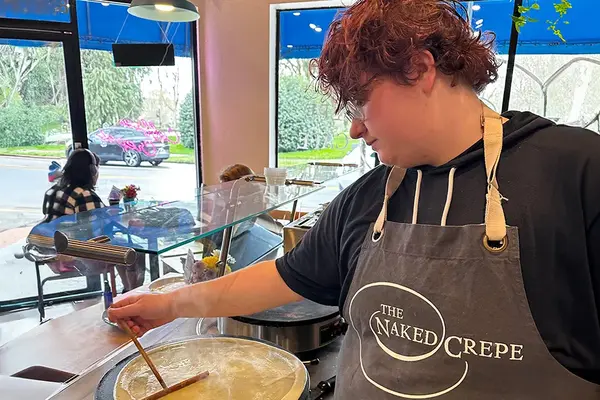 Sam Wade, wearing a grey apron embroidered with a cafe logo and the words "The Naked Crepe", stirs a pot of batter