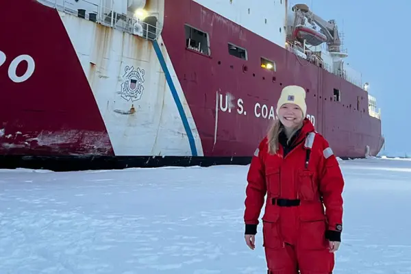 A woman in cold weather gear poses in a snowy landscape in front of a research vessel.