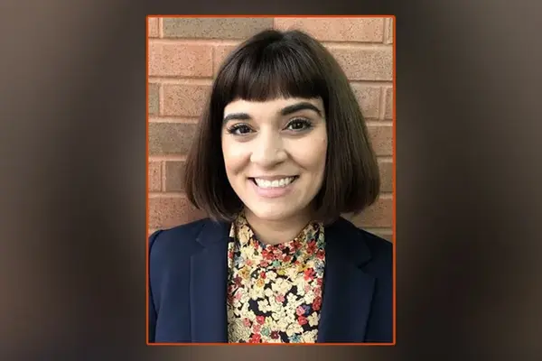 Alyssa Fuentez, wearing a floral blouse and blue jacket, smiles for the camera, posing in front of a brick background