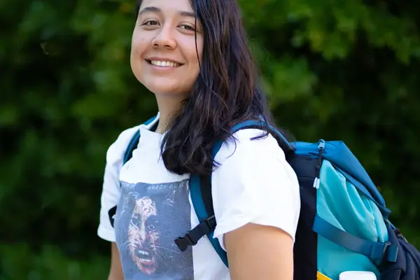 Celine Milam, wearing a T-shirt and a backpack, poses outdoors with green trees behind her
