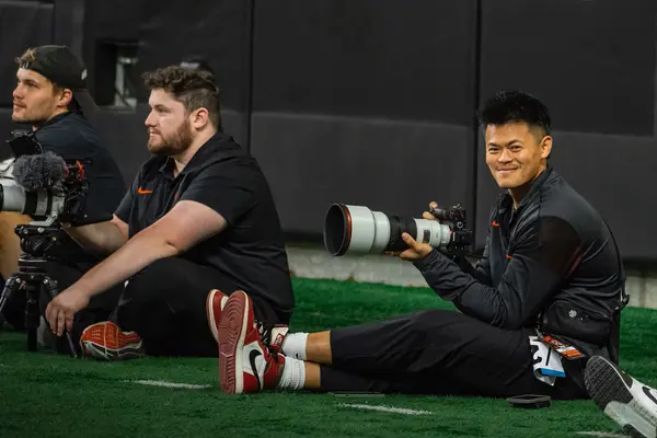 Nick Lin holds a camera with a zoom lens while sitting on the sidelines at a football field.