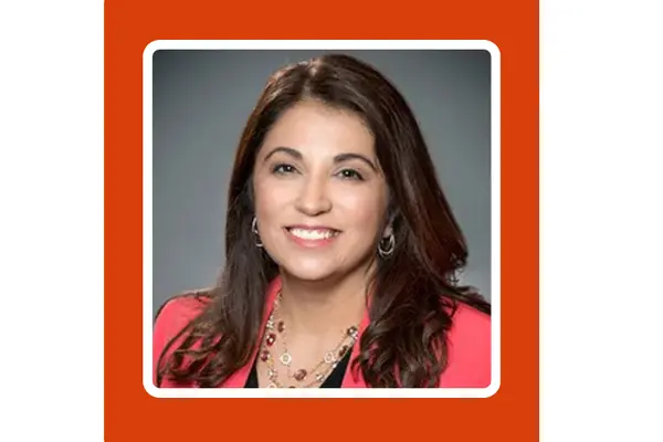 Veronica Garcia, wearing a coral-colored blazer, smiles for the camera in front of a neutral grey background