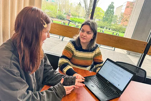 Two students have a discussion at a conference table while a laptop open to an AI chat screen is next to them.